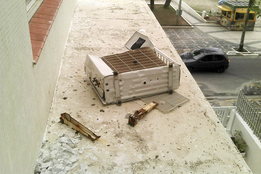Unidade condensadora de aparelho de ar condicionado e suportes enferrujados caídos em marquise de condomínio
