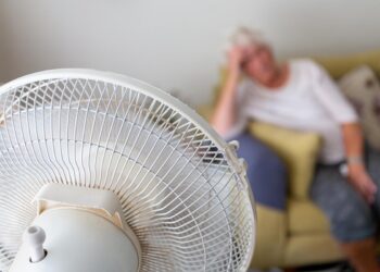 Mulher idosa em um sofá sofrendo de calor excessivo durante uma onda de calor e sendo resfriada por um ventilador elétrico oscilante em primeiro plano | Foto: Shutterstock