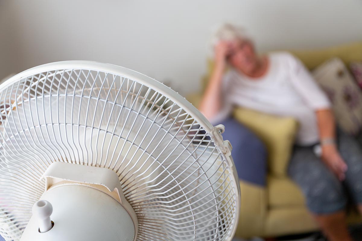 Mulher idosa em um sofá sofrendo de calor excessivo durante uma onda de calor e sendo resfriada por um ventilador elétrico oscilante em primeiro plano | Foto: Shutterstock